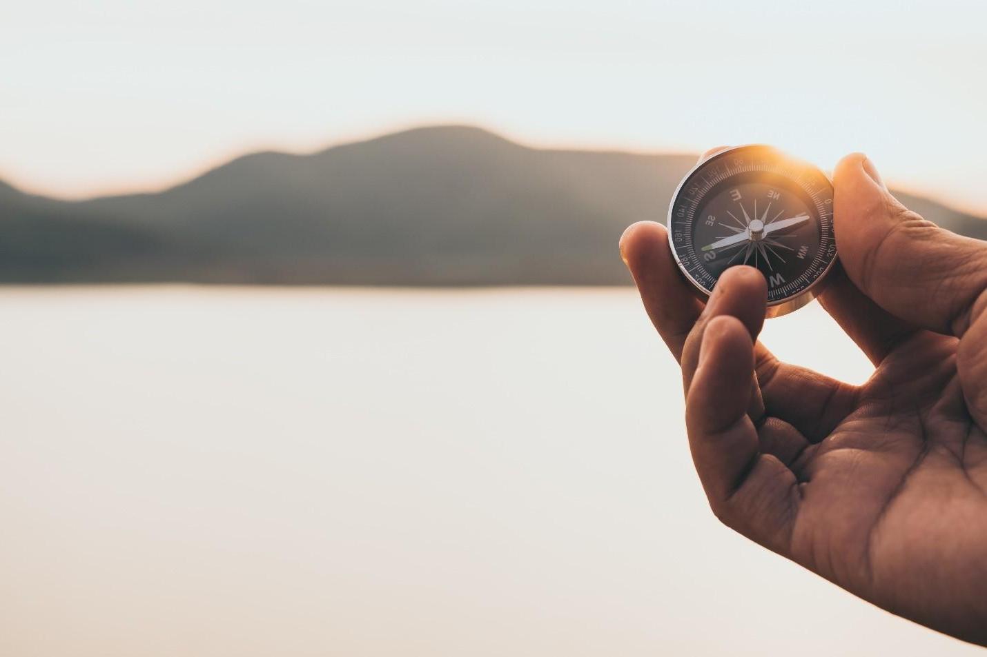 A hand holds a compass facing mountains and a body of water.